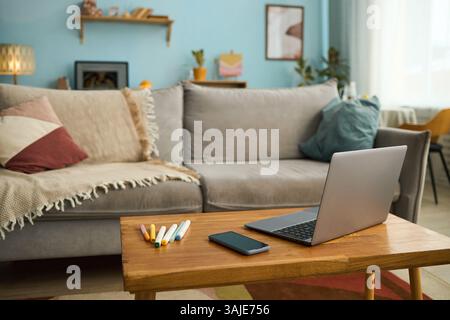 Salon moderne avec canapé confortable, table basse en bois et ordinateur portable, smartphone, crayons colorés placés sur la table créant une atmosphère chaleureuse Banque D'Images