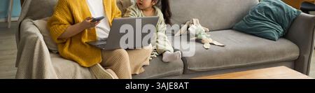 Parent en cardigan lumineux assis sur un canapé travaillant sur un ordinateur portable tandis que l'enfant en pyjama regarde attentivement Banque D'Images