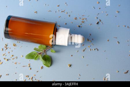 Bouteille d'huile d'origan affichée sur un fond bleu vif avec des feuilles d'origan fraîches à proximité. Banque D'Images