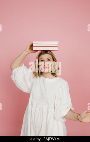 Femme en robe blanche équilibrant des livres sur sa tête de manière ludique. Banque D'Images