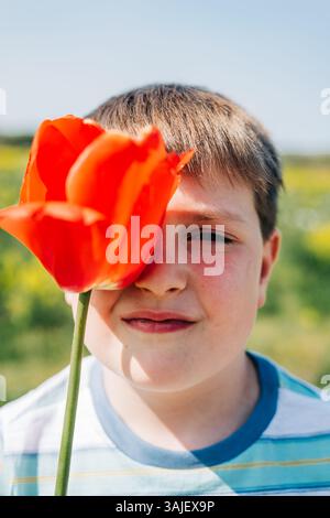 Garçon tient ludique tulipe rouge sur un œil dans la lumière du soleil Banque D'Images