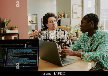 Plan moyen de développeurs biraciaux discutant du nouveau projet à la table en bois avec des ordinateurs portables sur le dessus dans le bureau moderne de la société INFORMATIQUE Banque D'Images