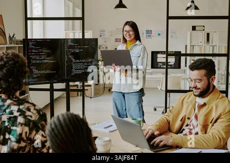 Programmeur souriant féminin asiatique délicat avec ordinateur portable dans les mains donnant la présentation, parlant de nouveaux conseils dans le codage devant le public dans le bureau de la société INFORMATIQUE Banque D'Images