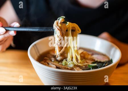 Une personne tenant des baguettes avec des nouilles et de la viande d'un bol de ramen, levant la vapeur, sur une table en bois. Banque D'Images