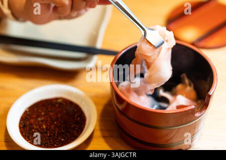 Une main tenant une fourchette avec de fines tranches de viande au-dessus d'un petit bol en bois. Le bol contient une sauce trempette, et il y a des baguettes et une assiette en t Banque D'Images