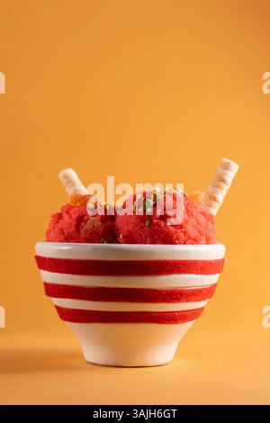 Crème glacée aux fraises servie dans un bol en céramique rayé avec fruits confits, pistaches et gaufrettes sur une table en bois. Gros plan sur le dessert d'été dans la rouille Banque D'Images