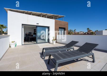 Espace terrasse extérieur avec deux chaises longues sombres et des plantes en pot, menant à un bureau à domicile lumineux et moderne via des portes coulissantes en verre. Banque D'Images