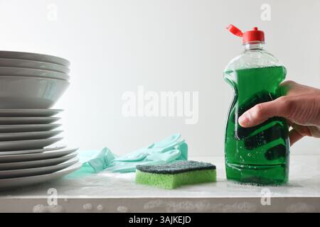 Bouteille tenant la main avec le produit vaisselle sur banc blanc avec de l'eau et de la mousse avec tampon à récurer et gants pour laver et empiler la vaisselle sur blanc Banque D'Images