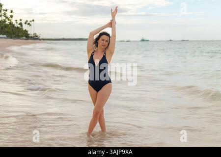 Une femme se tient gracieusement dans de douces vagues à la plage, mettant en vedette un maillot de bain noir alors qu'elle pose avec un bras levé. L'atmosphère est sereine avec un Banque D'Images
