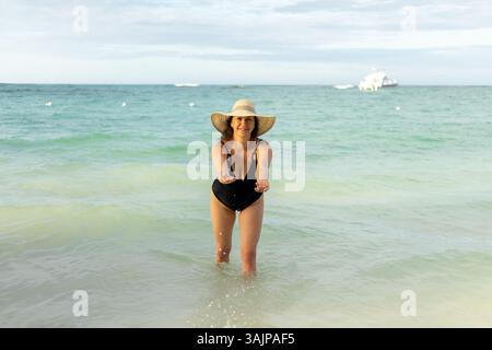 Une femme dans un maillot de bain noir se tient dans l'eau peu profonde, tendant la main avec les deux mains et souriant. La plage s'étend derrière elle comme le soleil se couche, créatine Banque D'Images