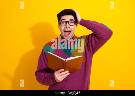 Jeune homme excité avec des lunettes tenant un livre sur un fond jaune tout en exprimant la surprise. Banque D'Images
