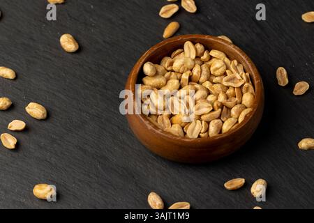 cacahuètes dans un bol rond en bois, cacahuètes croquantes sans coquilles sur une planche d'ardoise noire, vue de dessus Banque D'Images