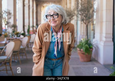 Femme caucasienne senior élégante dans le manteau et les lunettes debout dans la zone de café en plein air Banque D'Images