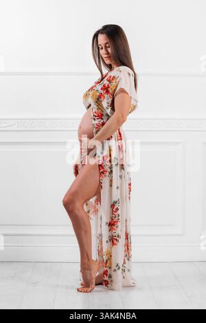 Mannequin enceinte portant une robe fleurie, posant dans un studio et touchant son ventre Banque D'Images