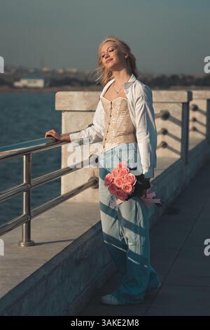 oman sur le pont, Une jeune femme aux longs cheveux blonds ondulés souriant à la caméra. Elle tient un bouquet de roses roses nouées avec un ribbo rose doux Banque D'Images