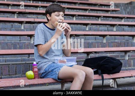 Manger sandwich, garçon assis sur des gradins avec boîte à lunch, pomme et sac à dos Banque D'Images