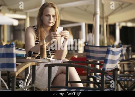 Femme attrayante se relaxant dans un transat au soleil en envoyant un sms ou en lisant des nouvelles sur son téléphone portable Banque D'Images