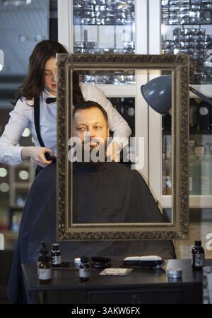 Homme d'âge moyen se faisant couper la barbe et les cheveux dans un salon de coiffure haut de gamme par une jeune femme en noeud papillon Banque D'Images