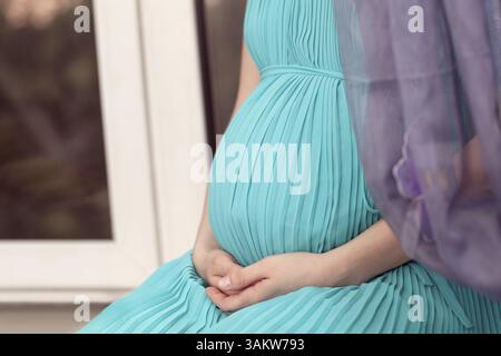 Close up of a pregnant woman wearing long robe bleue est assise sur le rebord de la fenêtre, le visage est méconnaissable Banque D'Images