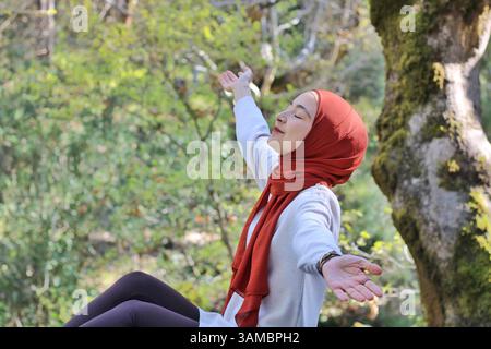 Femme avec une écharpe aux tons cannelle debout avec les bras ouverts et les yeux fermés, profitant d'un moment paisible entouré par la nature. Banque D'Images