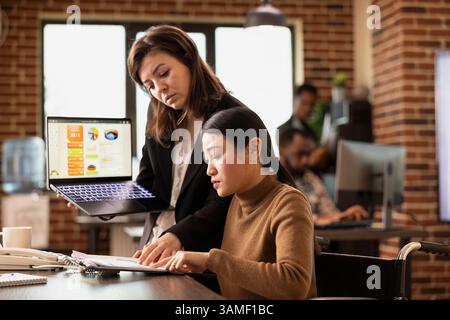 L'employée asiatique au bureau participe aux discussions d'affaires tout en gérant sa récupération dans un bureau moderne. Femme d'affaires se tient avec ordinateur portable, présentant des idées de marketing à un collègue en fauteuil roulant. Banque D'Images