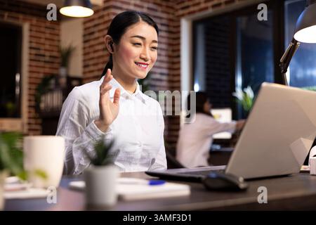 Gestionnaire de femme souriante agitant à l'écran de l'ordinateur portable, assistant à l'appel de vidéoconférence avec le client potentiel. Femme entrepreneure asiatique utilisant son ordinateur personnel pour une réunion virtuelle avec des partenaires commerciaux. Banque D'Images