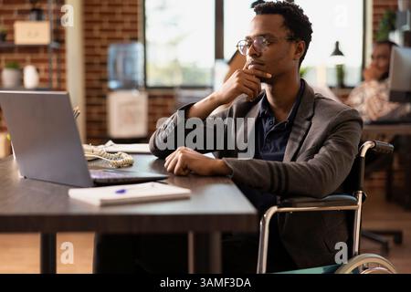 Homme d'affaires noir en fauteuil roulant tenant son menton et se concentrant sur les données financières sur son ordinateur portable. Employé masculin avec un handicap, axé sur l'examen d'un plan de projet d'affaires dans le bureau de démarrage. Banque D'Images