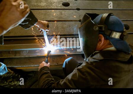 Un soudeur, tenant la tête de soudage automatique, et un assistant, serrant fermement les pièces métalliques ensemble, effectuent le processus de soudage automatisé sur un travail Banque D'Images