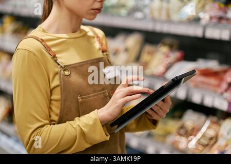 Employé du magasin dans un tablier tenant la tablette, tout en vérifiant l'inventaire dans l'allée de l'épicerie remplie de divers produits placés sur les étagères portant une chemise jaune axée sur la tâche Banque D'Images