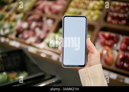 Main humaine tenant smartphone dans l'épicerie avec des produits frais affichés sur les étagères en arrière-plan. L'écran vide du smartphone peut être utilisé pour ajouter des dessins ou du texte Banque D'Images