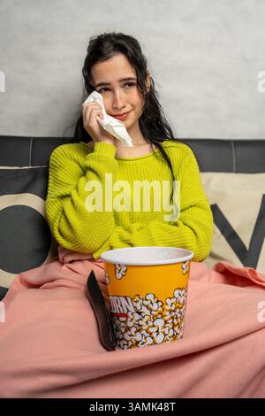 Jeune femme essuyant les larmes avec du tissu tout en regardant un film triste à la télévision, appréciant le pop-corn sur le canapé Banque D'Images