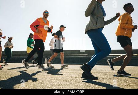 Un coureur masculin, portant une veste orange vif et des lunettes de soleil, fait des gestes ludiques au pistolet tout en participant à une course sur route communautaire animée. Banque D'Images