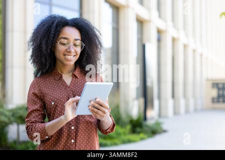 Une jeune femme avec des lunettes sourit alors qu'elle utilise une tablette à l'extérieur près d'un bâtiment moderne. Elle porte une chemise élégante à motifs. Banque D'Images