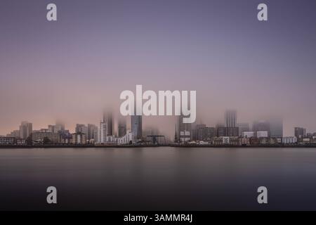 Canary Wharf tourné lors d'une journée brumeuse au coucher du soleil, permettant aux couleurs du ciel de se diffuser et d'envelopper le paysage urbain à l'heure dorée. Banque D'Images