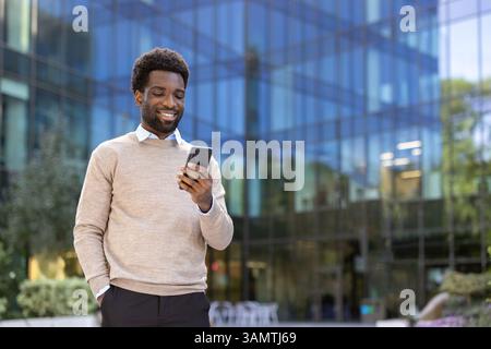 Un homme noir attrayant sourit en regardant son smartphone, debout à l'extérieur devant un immeuble de bureaux moderne. Il semble heureux. Banque D'Images