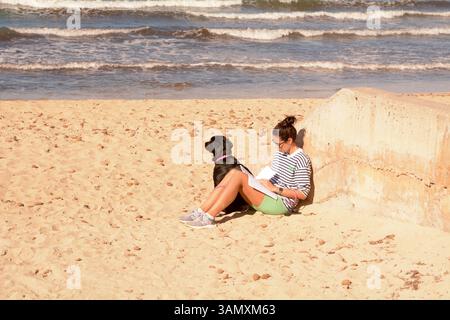 Une femme est assise lisant sur la plage des Meravelles tandis que son animal de compagnie labrador donne sur la baie de Palma, île de Majorque, en Espagne Banque D'Images