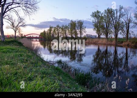 Vue sereine sur la rivière avec une surface d'eau calme montrant des reflets clairs, bordée d'arbres sous un ciel paisible du soir, avec une arche distinctive Banque D'Images