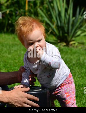 Les mains des pères aident à tenir une fille de bébé de cheveux rouges dans une cour arrière. Jeune fille jouant avec un seau à l'extérieur en été. Banque D'Images