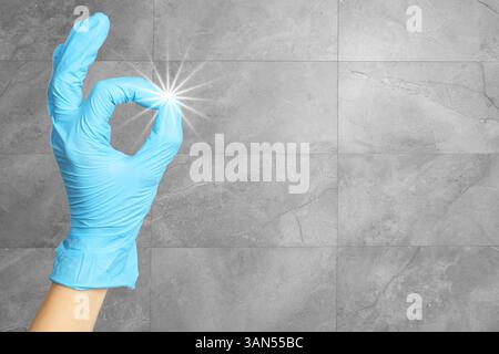 Un employé du service de nettoyage portant des gants en caoutchouc montre un geste correct près d'un mur carrelé gris, gros plan. Brillance de pureté sur le doigt Banque D'Images