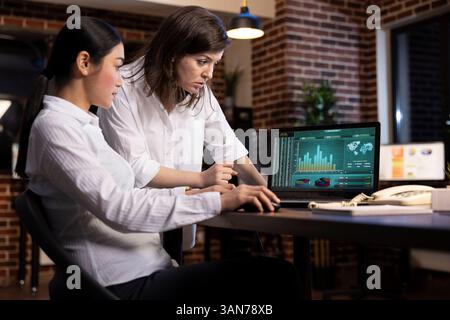 Une discussion professionnelle entre collègues, collaborant à un projet d'entreprise, utilisant des données de recherche sur ordinateur portable. Femme blanche se lève et examine les détails tandis que ses collègues font défiler le document. Banque D'Images