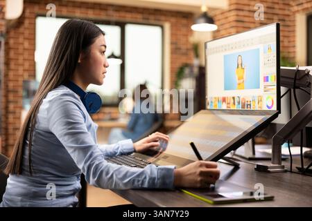 Illustrateur professionnel féminin utilisant le stylet sur la tablette graphique, créant un croquis numérique. Femme asiatique travaillant dans l'espace de travail de l'agence créative, axée sur l'édition et la conception de contenu pour la production. Banque D'Images