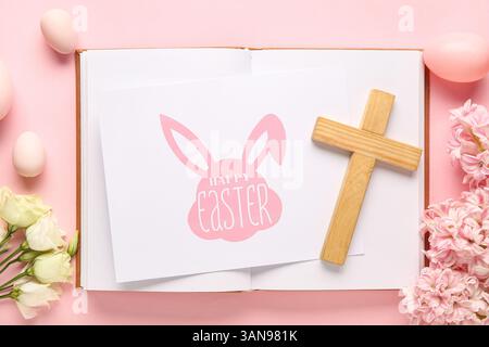 Croix en bois avec livre, oeufs, fleurs et carte de voeux JOYEUSES PÂQUES sur fond rose Banque D'Images