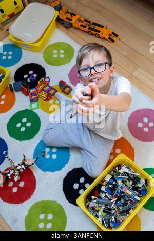 Garçon heureux jouant avec des blocs colorés à la maison. Garçon souriant jouissant d'un jeu créatif avec des blocs colorés et des jouets sur tapis à motifs dans une chambre confortable. Banque D'Images