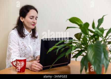 Femme travaillant sur ordinateur portable avec du café et des plantes. Une jeune femme dans une blouse blanche est assise à un bureau à l'aide d'un ordinateur portable, avec une tasse à café rouge et une plante verte Banque D'Images