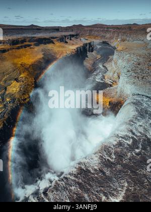Vue aérienne de la majestueuse cascade Dettifoss avec un arc-en-ciel vibrant au milieu de falaises accidentées et de brume puissante, Vatnajokull, Islande. Banque D'Images