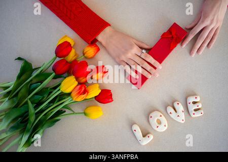 Tulipes, boîte-cadeau et biscuits sur le thème de l'amour. Un gros plan de mains près d'une boîte cadeau rouge, tulipes colorées et biscuits orthographiant « AMOUR », symbolisant la romance Banque D'Images