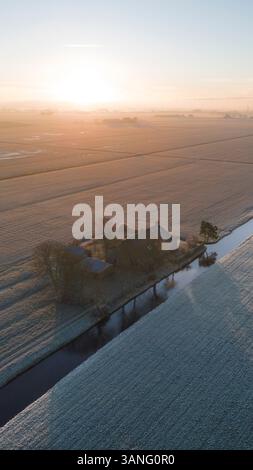 Vue aérienne d'une ferme paisible avec une maison entourée d'une prairie enneigée au lever du soleil, Sneek, pays-Bas. Banque D'Images