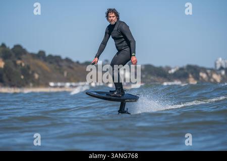 Un homme dans une combinaison noire monte une tranchée hydrofoil électrique foilboard, dans la mer par une journée ensoleillée, efoil, foil drive, foil hybride assistance. Banque D'Images