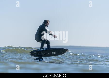 Un homme dans une combinaison noire monte une tranchée hydrofoil électrique foilboard, dans la mer par une journée ensoleillée, efoil, foil drive, foil hybride assistance. Banque D'Images