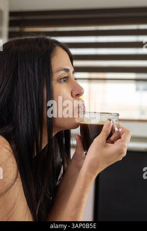 Une jeune femme souffle doucement sur sa tasse à café, attendant qu'elle refroidisse, alors qu'elle profite d'un moment calme et réfléchi dans un cadre confortable. Banque D'Images